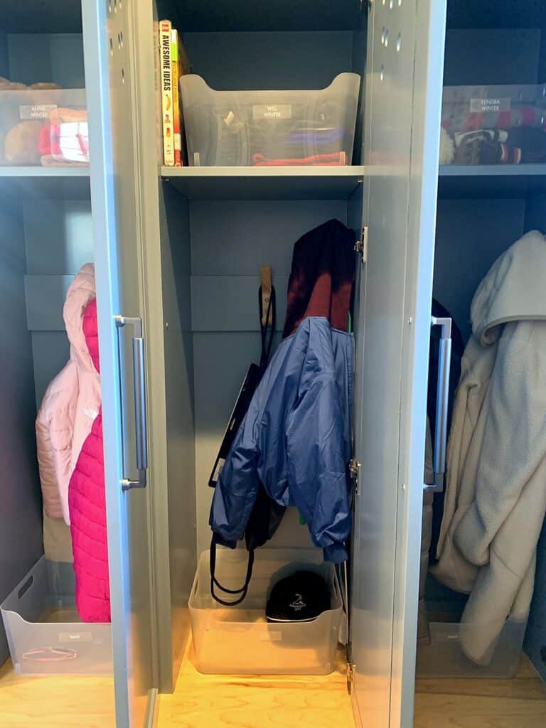 Kids' jackets hanging on hooks inside a mudroom locker with a clear bin below