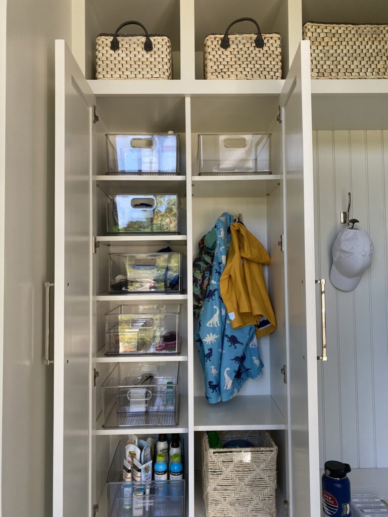 Mudroom locker organized with stacked clear bins, a raincoat on a hook, and baskets up top