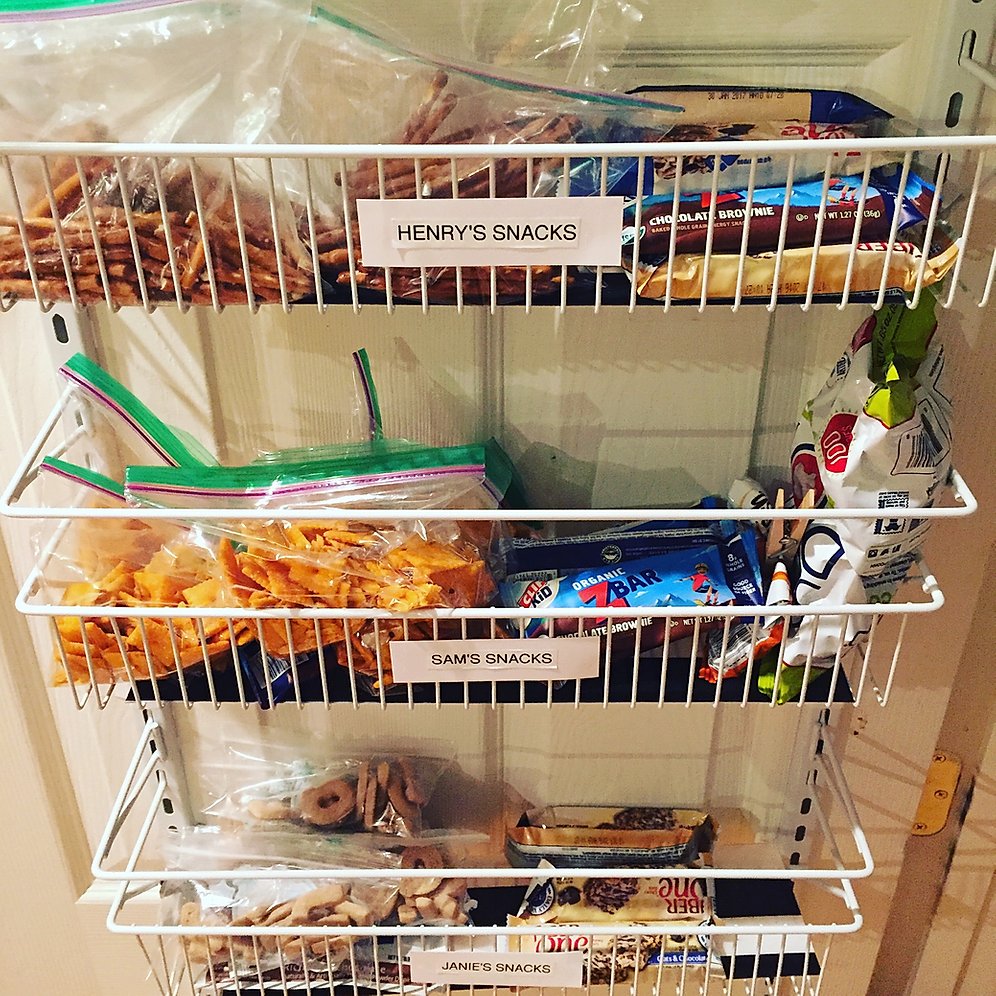 Pantry door rack with baskets holding separate snacks labeled with different kid's names for easy snack grabs for school lunch. 