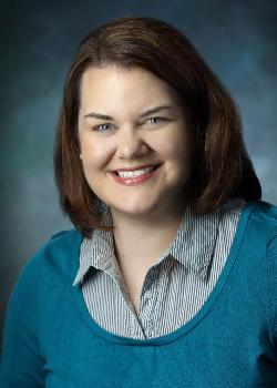 Photo of Kristen Lindeman Shifflett. Caucasian woman with green eyes and shoulder length brunette hair, smiling wearing a striped collared shirt under a blue scoop necked sweater.