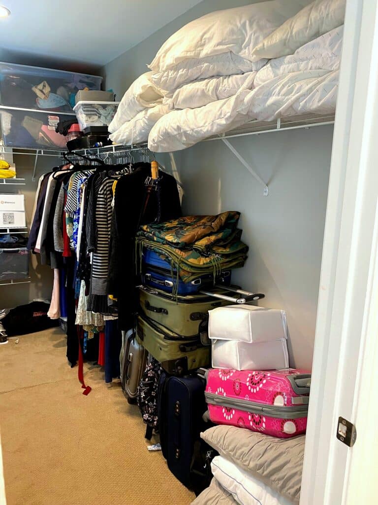 Cluttered walk-in closet with dense clothing rack, stacked suitcases, and heaped comforters before professional organization