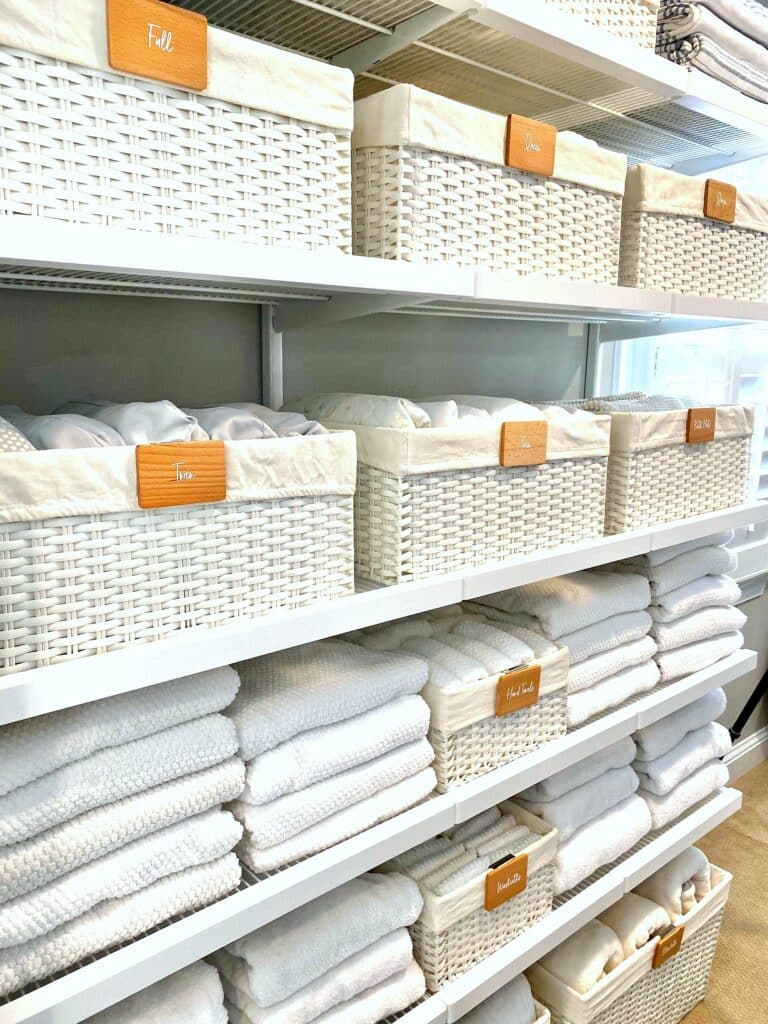 Beautifully organized linen wall with labeled white wicker baskets by bed size and neatly folded white towels after professional organization