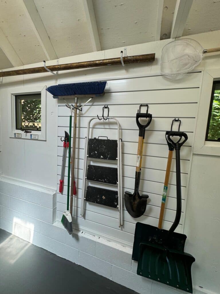 Garden tools, shovels, and outdoor equipment neatly organized on slat wall in white Annapolis shed