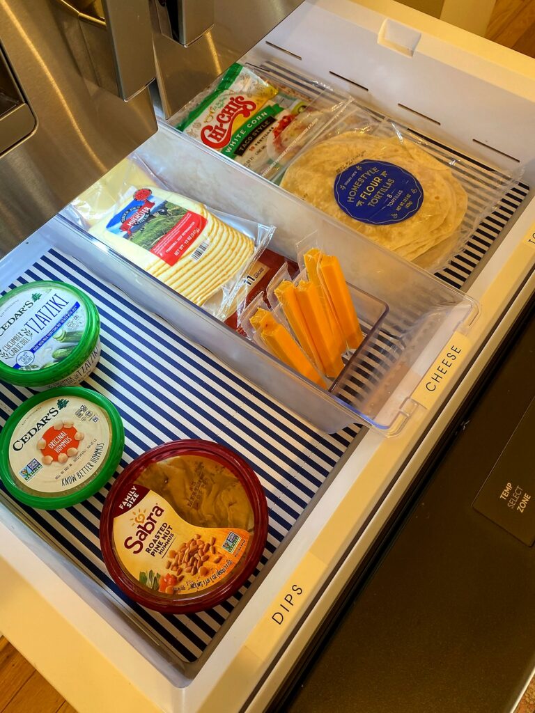 Refrigerator deli pull-out drawer with labeled dips and cheese zones holding hummus, tzatziki, and tortillas