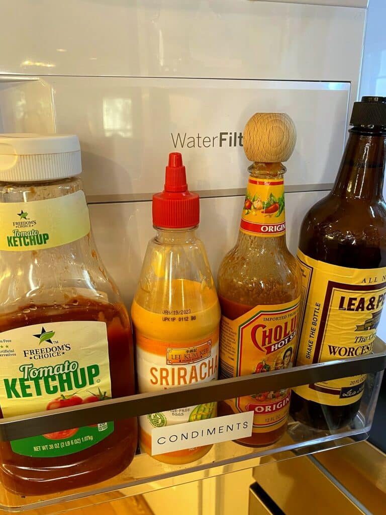 Refrigerator door bin labeled Condiments with ketchup, sriracha, Cholula, and Worcestershire sauce