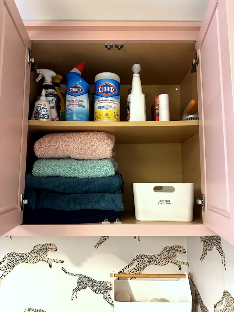 Laundry cabinet with cleaning sprays on the top shelf and folded towels beside a labeled bin