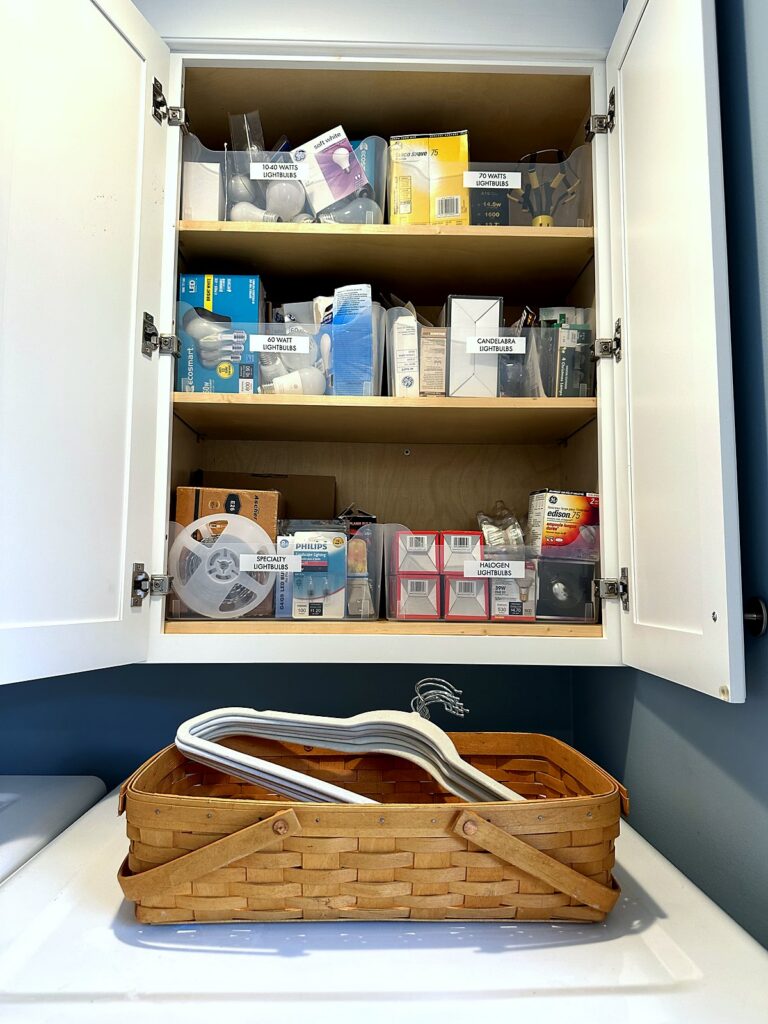 Labeled boxes organized in a laundry cabinet with a basket of hangers on the dryer below