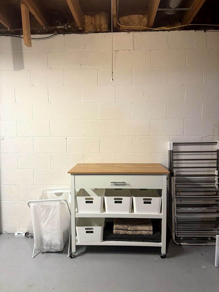 Rolling cart with labeled white bins and a slim hamper in a basement laundry area