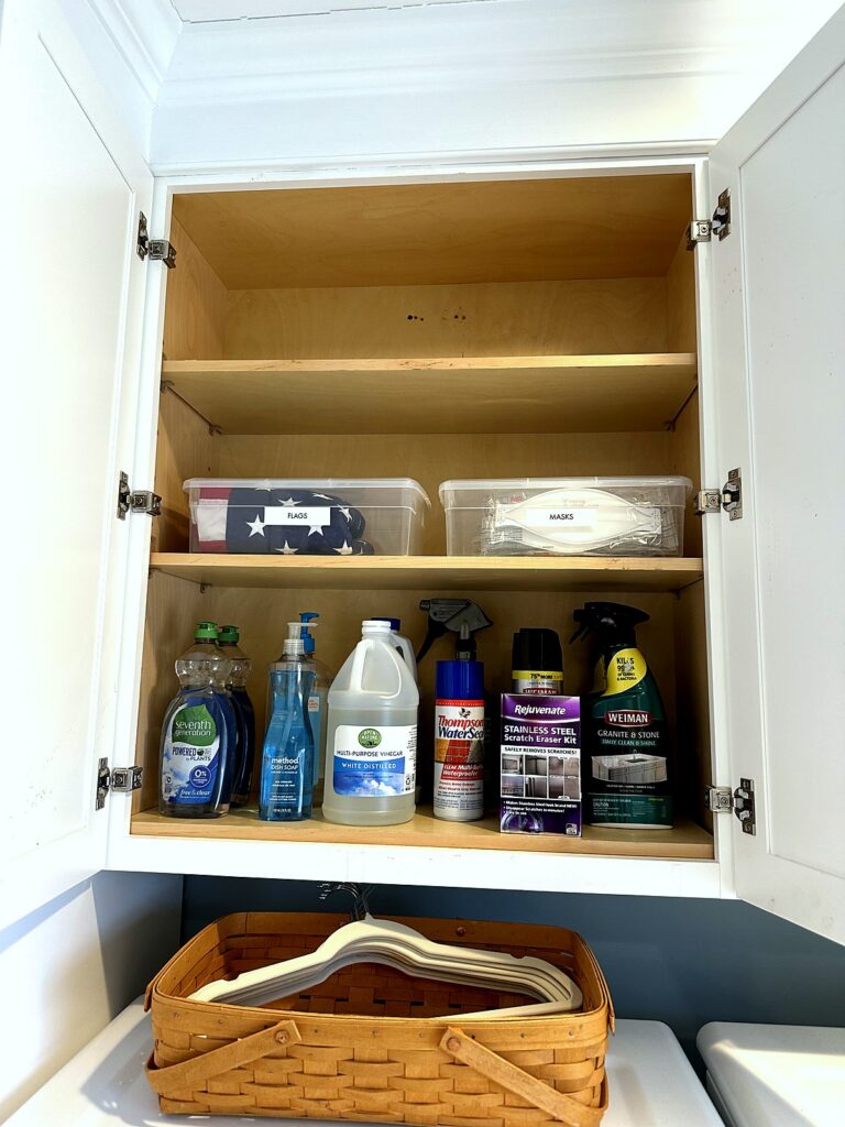 Labeled bins and grouped cleaning supplies in a laundry room cabinet above a basket of hangers
